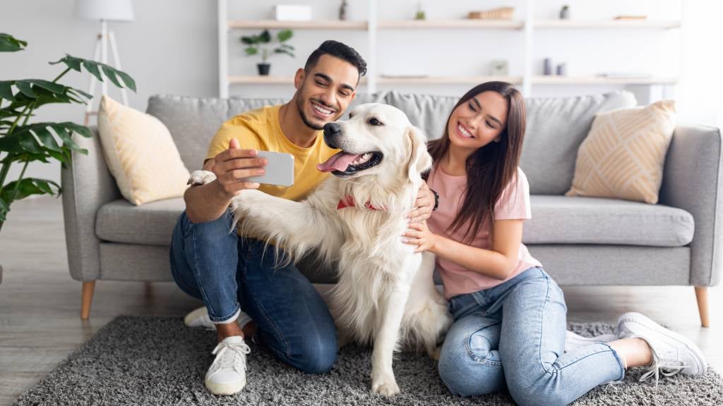 A happy couple taking a selfie with their dog on their living room floor.