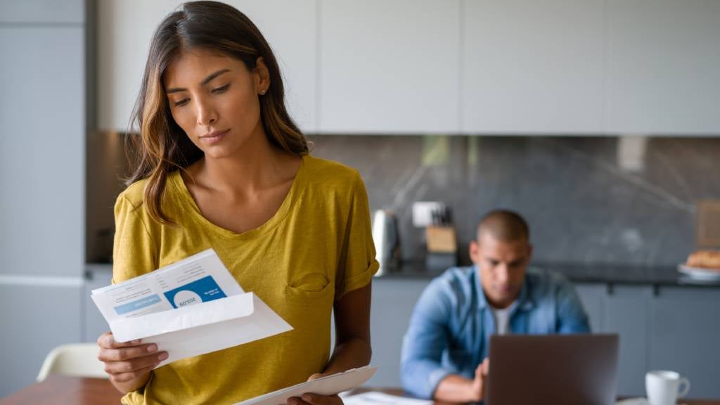 A worried woman looks at a bill and a man sits surrounded by bills at his laptop