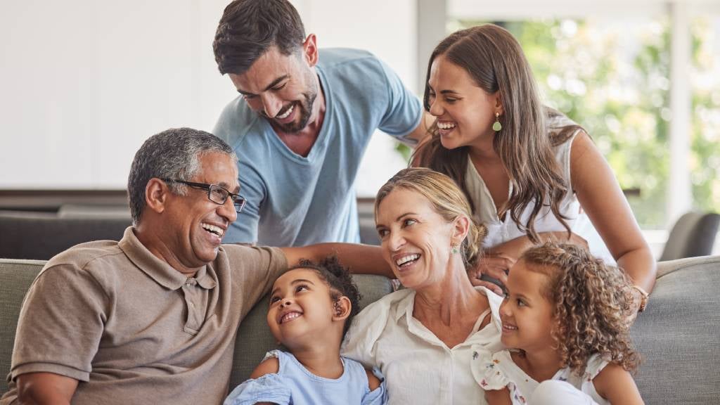 Grandparents sit on a sofa playing with their grandkids while the parents look on