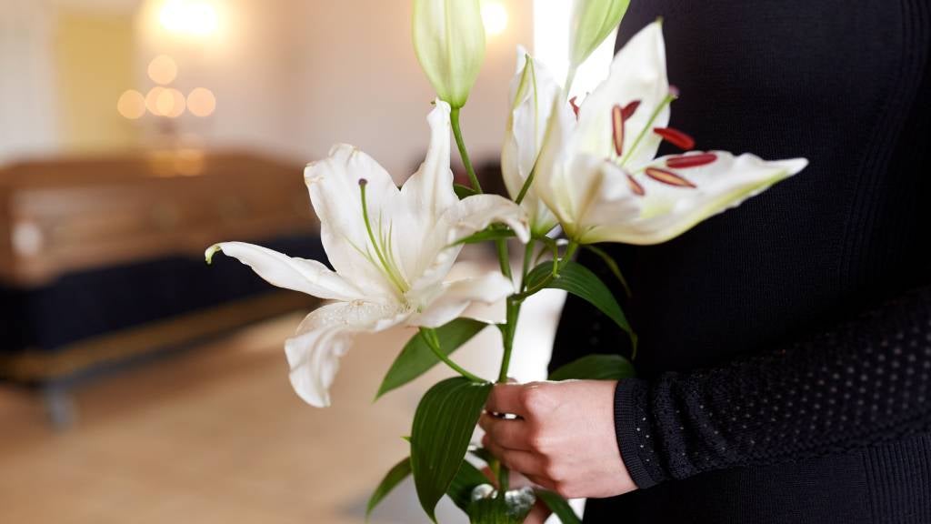 A woman holding lilies stands in front of a coffin