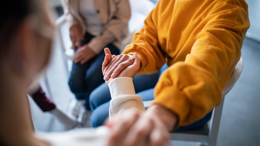 Women sit on chairs and one of them holds another’s hand in comfort