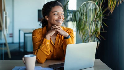 A content looking professional woman sits at her desk with a morning coffee