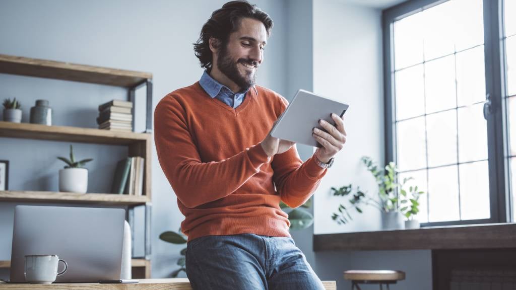 A young professional man leans on his desk and smiles at his tablet