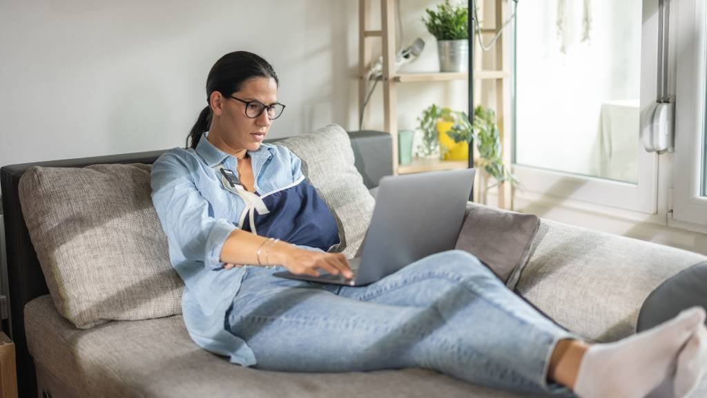 A woman with one arm in a brace reclines on a daybed and uses her laptop